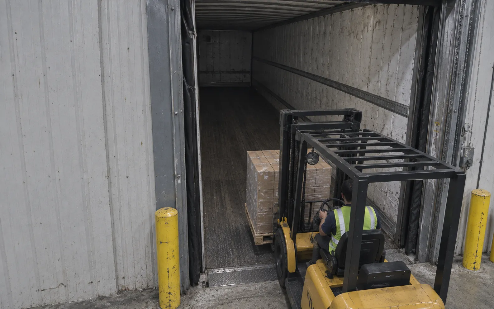 Building-mounted camera looking through the dock opening into the trailer, behind the forklift and its pallet load.