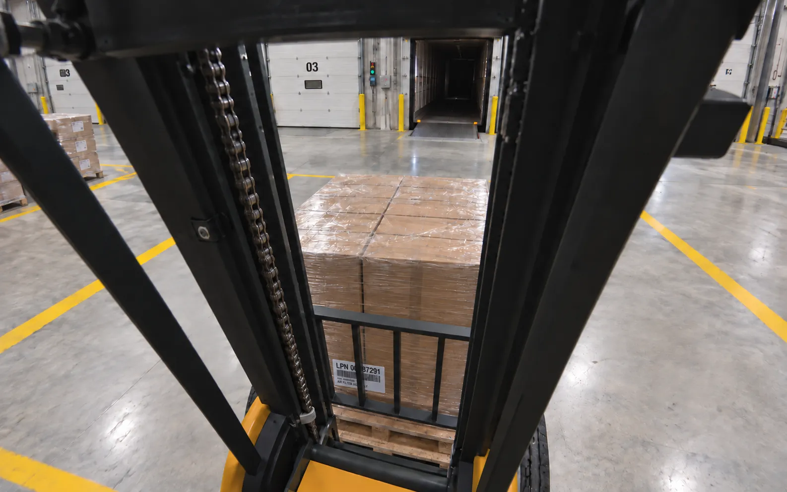 Camera inside the forklift overhead guard looking forward through the mast at a pallet carried on the forks toward the open loading dock.