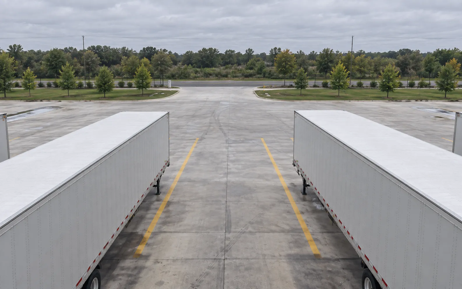 View from a PTZ mounted above the exterior dock doors, looking outward between parked trailers across the concrete yard toward the distant entrance and tree-lined boundary.