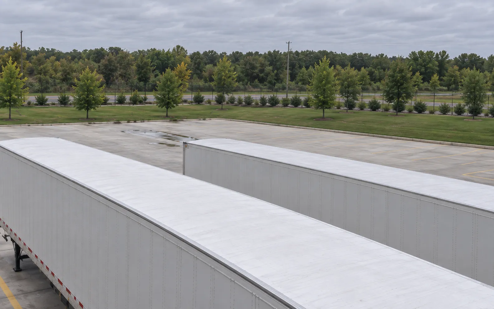 The same dock-wall PTZ panned across white trailer roofs toward the yard edge, grass verge, perimeter fence and distant trees.